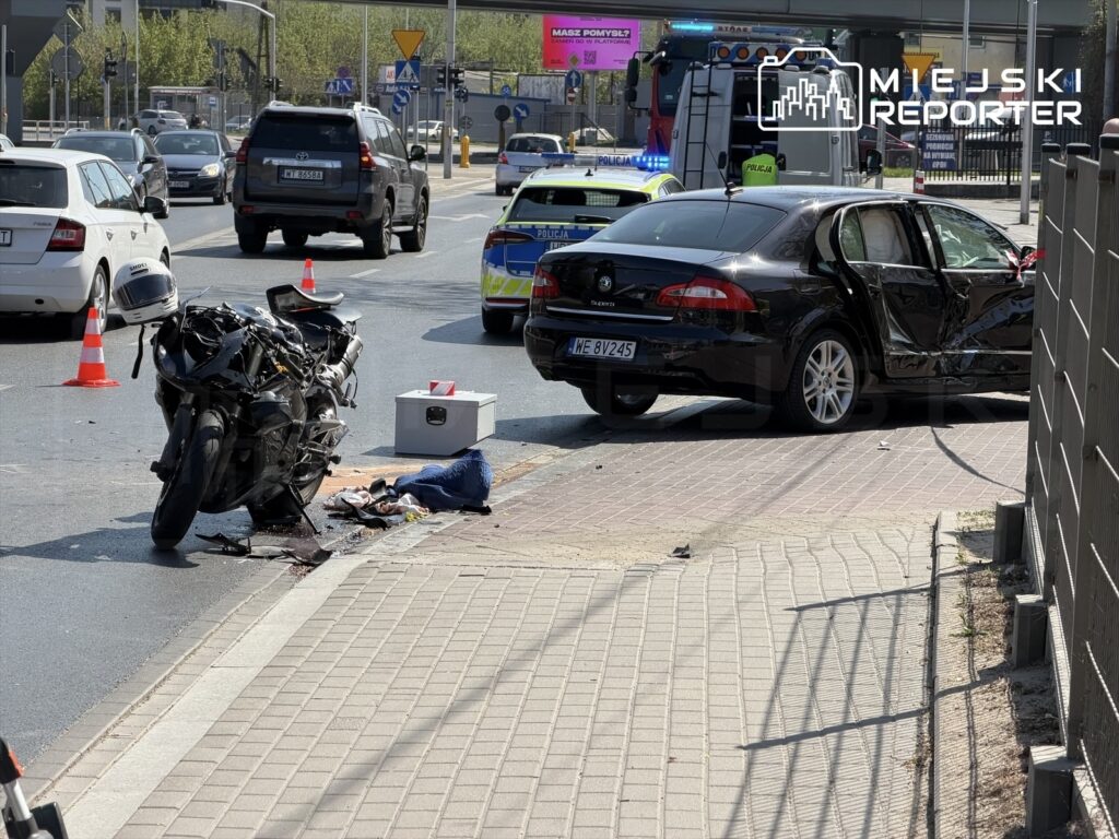 Motocykl leży na jezdni obok rozbitego czarnego samochodu osobowego, w tle widać pojazdy policyjne i sygnalizację drogową.