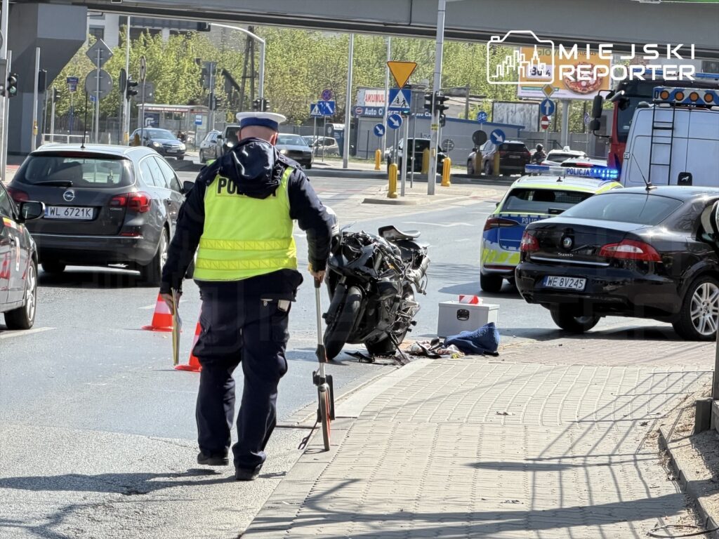 Policjant w kamizelce odblaskowej bada miejsce wypadku motocyklowego na ruchliwej ulicy, obok leży uszkodzony motocykl.
