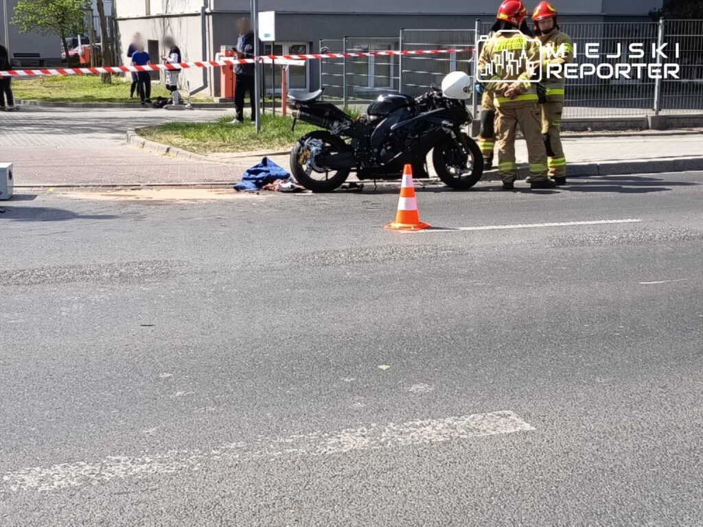 Dwaj strażacy w hełmach rozmawiają obok motocykla, oznaczonego pomarańczowym pachołkiem, na ulicy z barierkami.