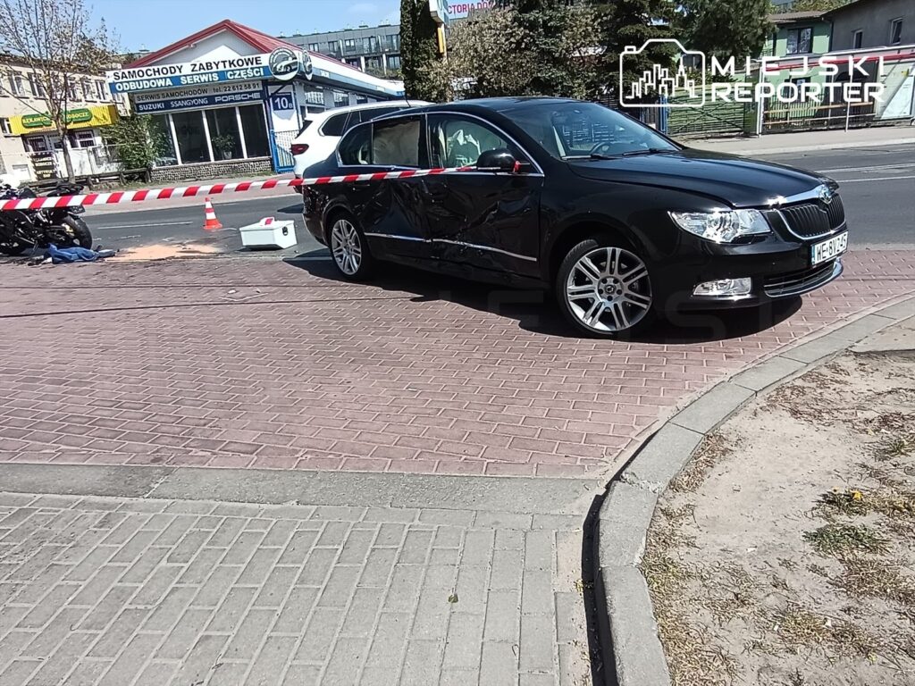 Czarny samochód osobowy z uszkodzeniem stoi na chodniku, obok zabezpieczony taśmą ostrzegawczą. W tle widoczny warsztat samochodowy.