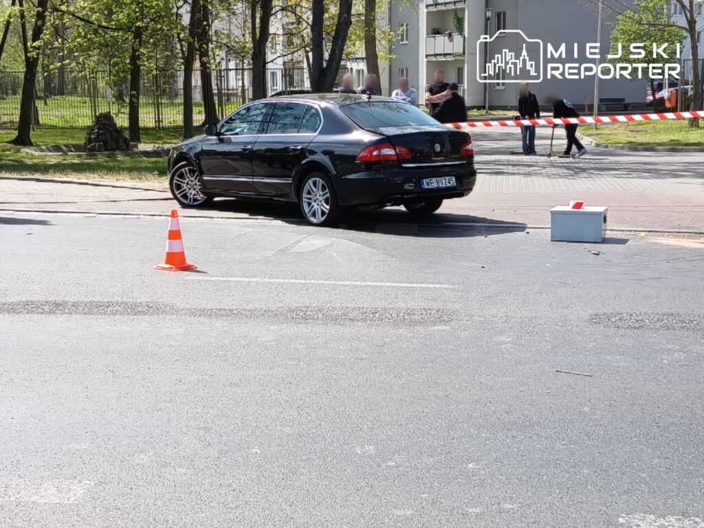 Czarny samochód osobowy zaparkowany na jezdni, w tle grupa osób oraz taśma zabezpieczająca miejsce zdarzenia.