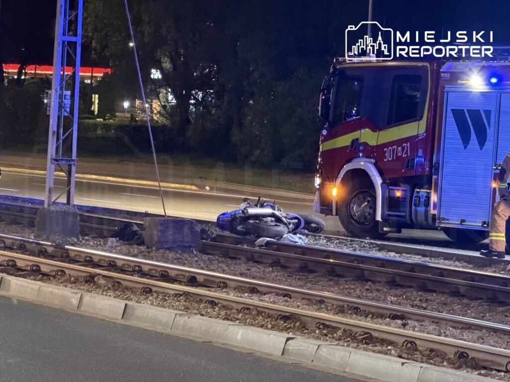 Na torach kolejowych leży przewrócony motocykl, obok stoi wóz strażacki z włączonymi sygnałami.