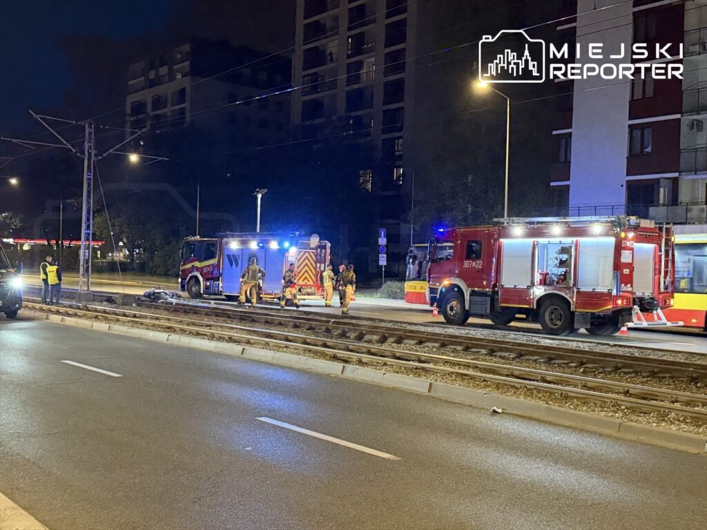 Grupa strażaków i ratowników medycznych pracuje przy torach tramwajowych w nocy, obok wozu strażackiego.