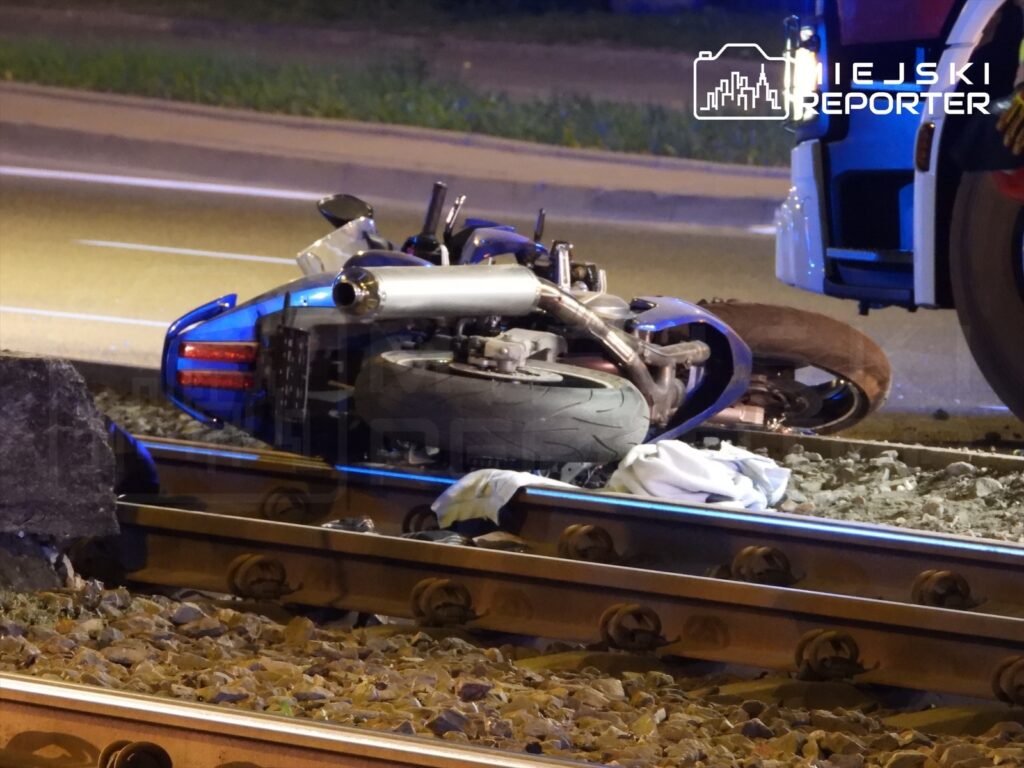 Motocykl leży na torach kolejowych, obok niego widoczna jest biała tkanina, a w tle widać pojazd służb ratunkowych.