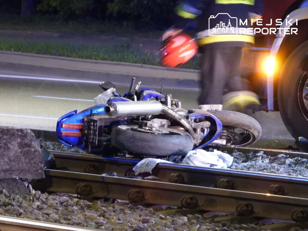 Motocykl leży na torach kolejowych, obok niego pracują strażacy w mundurach.