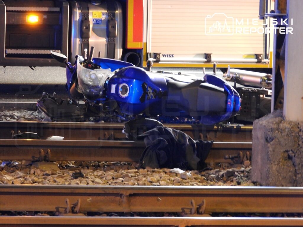 Nieprzewidziane zdarzenie drogowe, gdzie przewrócony niebieski motocykl leży na torach kolejowych obok fragmentów odzieży.