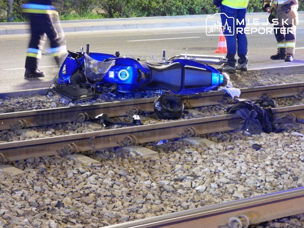 Motocykl leży na torach kolejowych, otoczony przez strażaków i ratowników, w nocy.