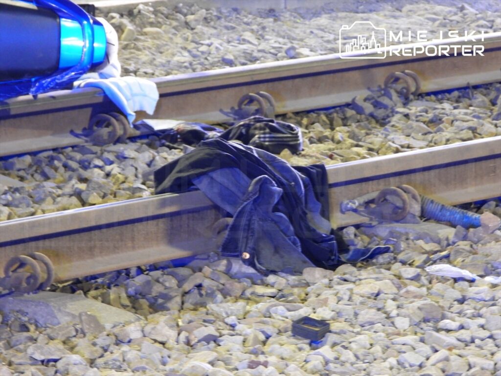 Na torach kolejowych leżą porzucone ubrania wśród kamieni, w tle widoczny jest fragment niebieskiego pojazdu.