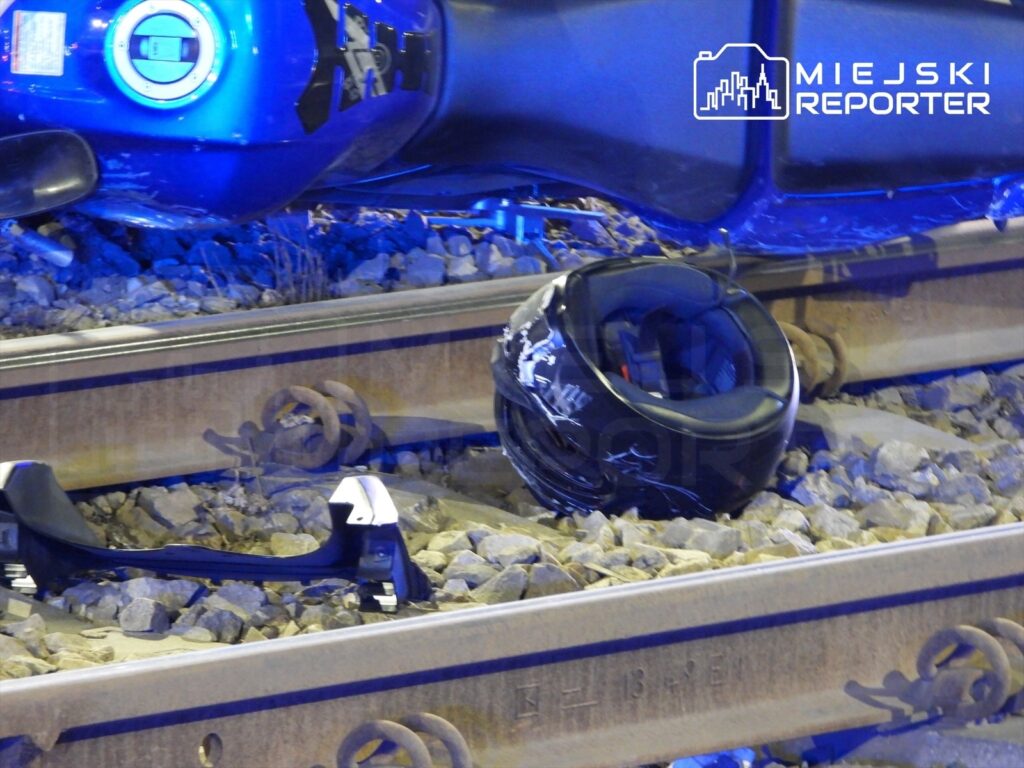 Na torach kolejowych leży czarny kask motocyklowy obok fragmentów uszkodzonego pojazdu.