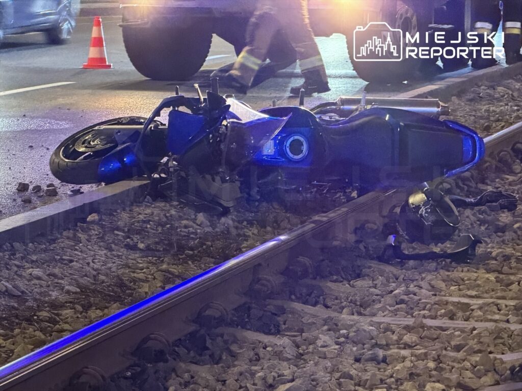 Motocykl leżący na torach kolejowych, obok widoczne fragmenty drogi oraz pachołki drogowe.