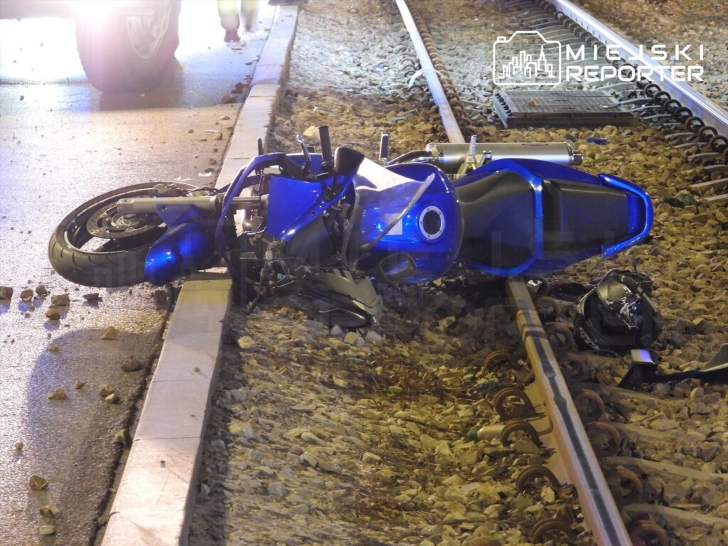 Motocykl w niebieskim kolorze leży na torach kolejowych, obok widoczne odłamki i kask.