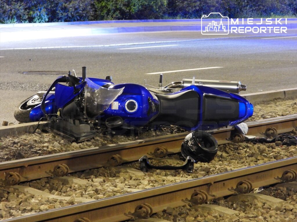 Złamana niebieska motocykl leży na torach tramwajowych obok jezdni, obok niego kask i fragmenty odzieży.