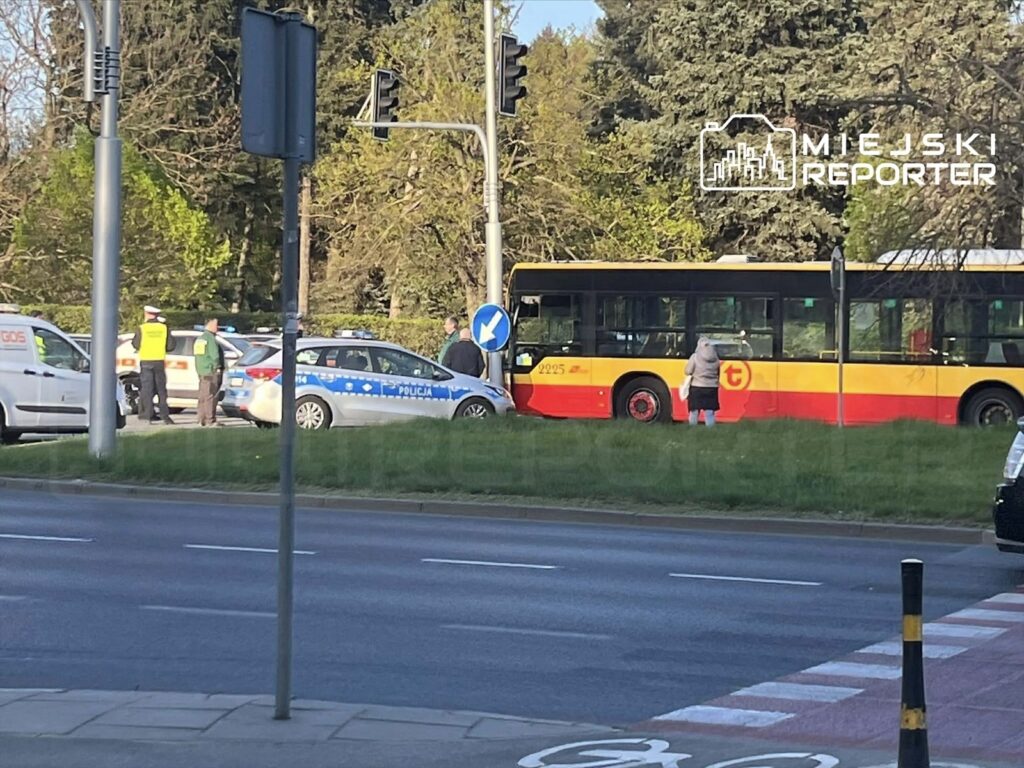 Grupa funkcjonariuszy policji i pracowników służb porządkowych prowadzi czynności na skrzyżowaniu, obok przystanku autobusowego.