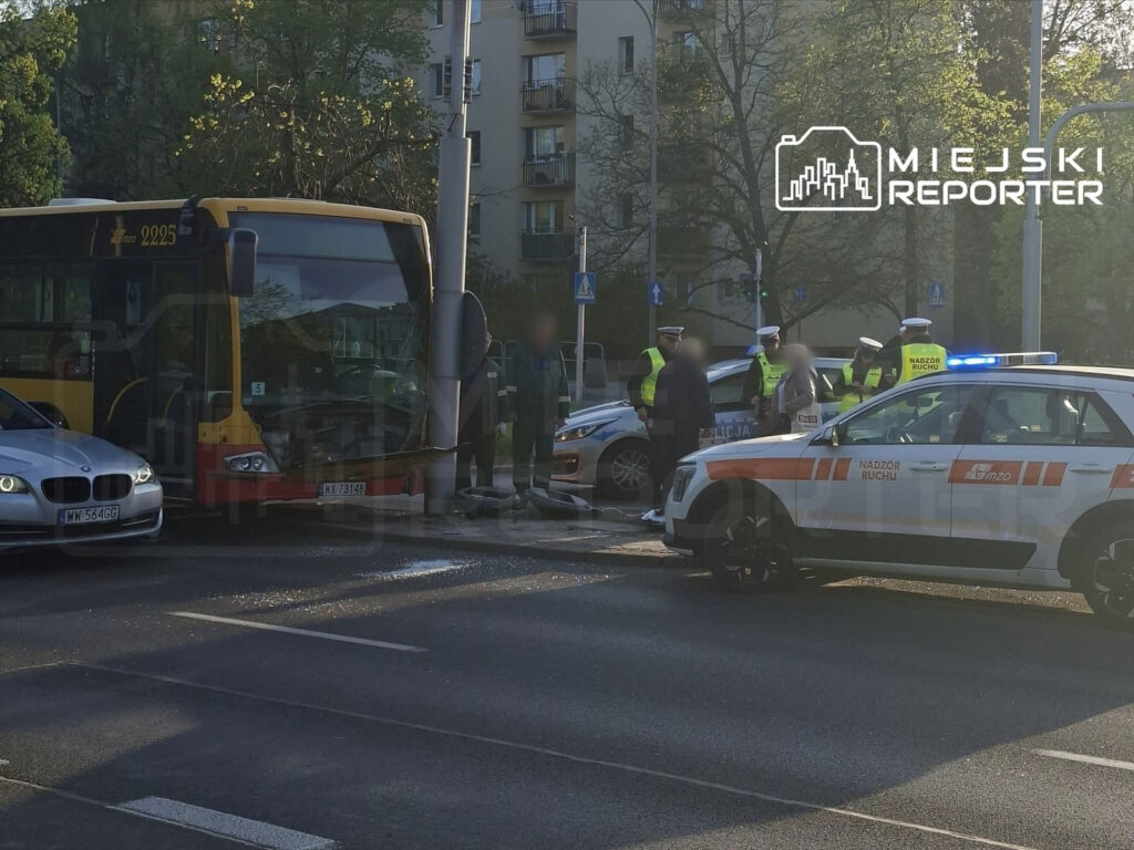 Zderzenie autobusu z latarnią, w tle funkcjonariusze policji rozmawiający z kierowcą osobówki.