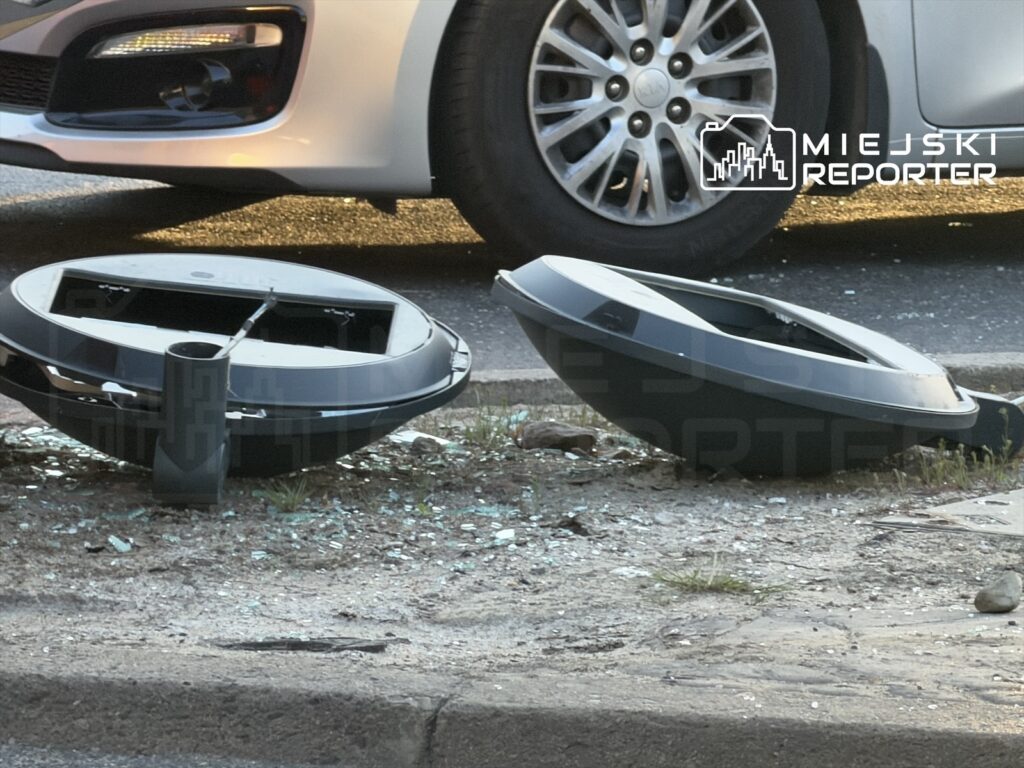 Dwa uszkodzone pojemniki na odpady leżą na chodniku obok zaparkowanego samochodu, w otoczeniu szkła i trawy.