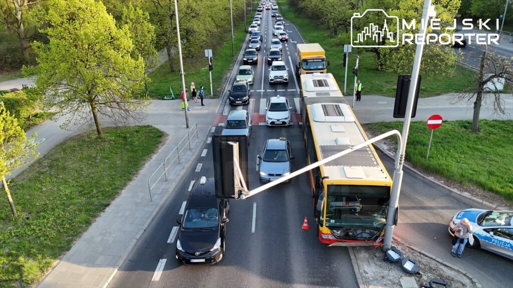 Ruch samochodowy zablokowany na ulicy, obok uszkodzony autobus i policyjny radiowóz, w tle drzewa i chodnik.