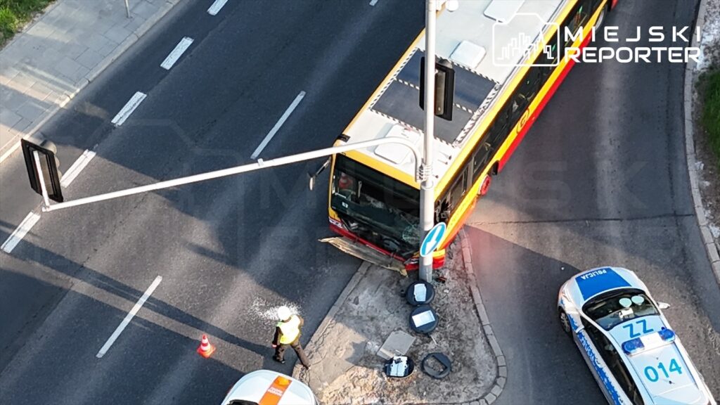 Autobus zatrzymany przy skrzyżowaniu, kierowca w kamizelce odblaskowej sprawdza uszkodzenia, obok policyjny radiowóz.