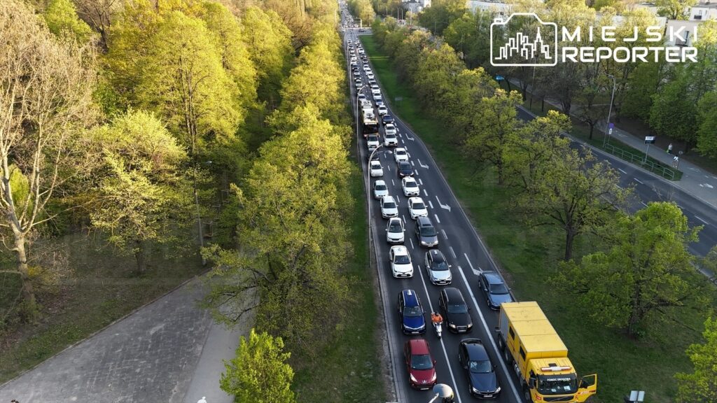 Ruch samochodowy wzdłuż drogi, z długim korkiem pośród zieleni drzew i nowoczesnych budynków.