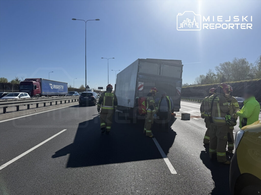 Grupa strażaków w mundurach omawia sytuację przy uszkodzonym samochodzie ciężarowym na drodze.