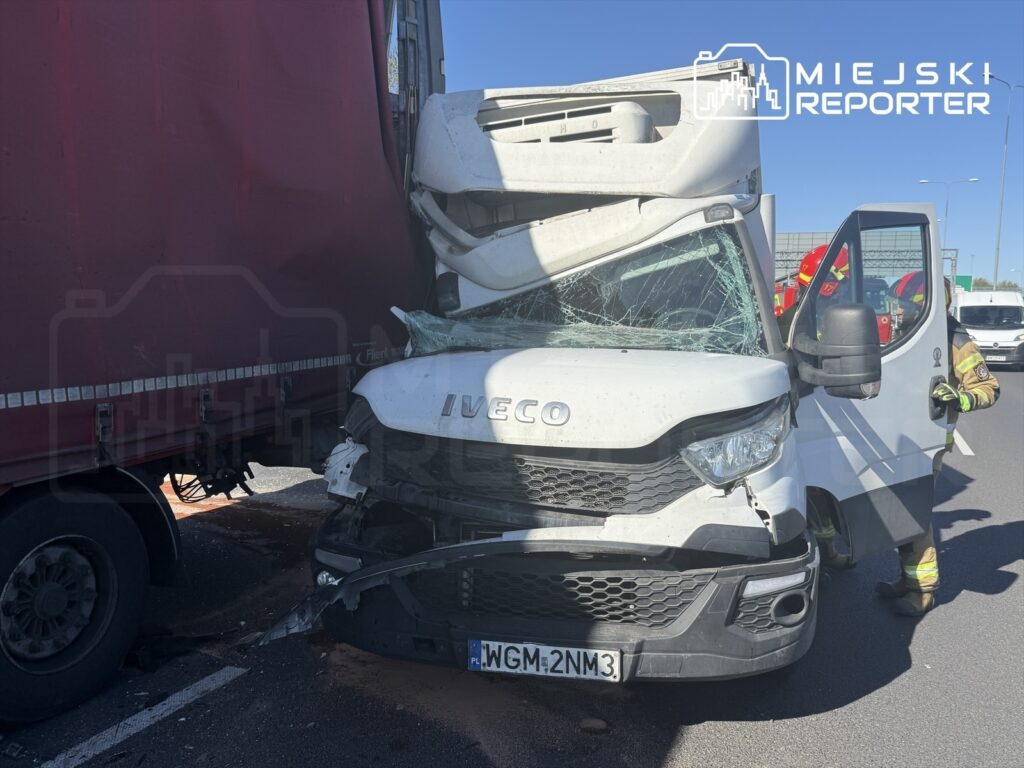 Zderzenie białego furgonu z czerwoną ciężarówką na drodze, z widocznymi uszkodzeniami pojazdów i rozbitym przednią szybą.