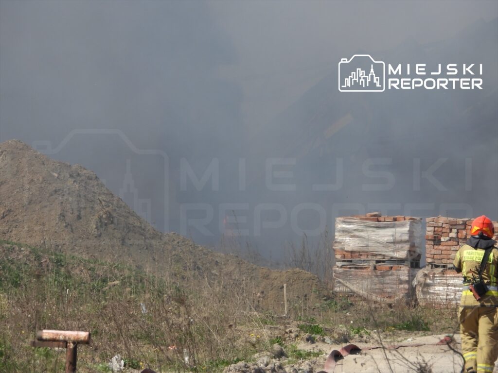 Strażak w hełmie i mundurze obserwuje dym unoszący się z placu budowy w otoczeniu gruzów i zarośli.