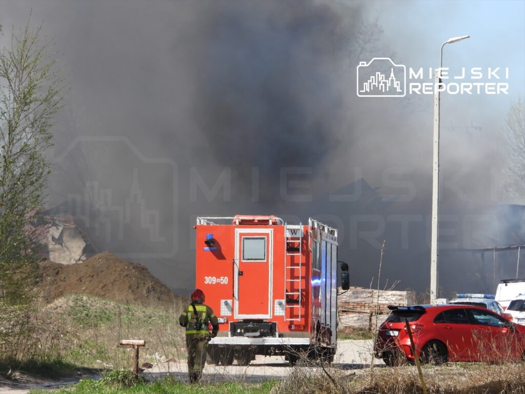 Strażak w mundurze podchodzi do czerwonego wozu strażackiego w pobliżu dużego pożaru, z którego unosi się dym.