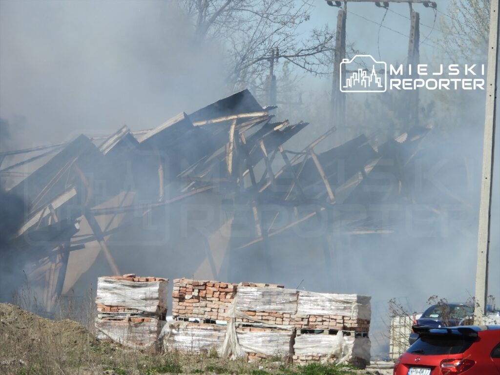Zniszczony budynek z dachem w opadzie, otoczony gęstym dymem, w tle widoczne palety z cegłami i samochód osobowy.