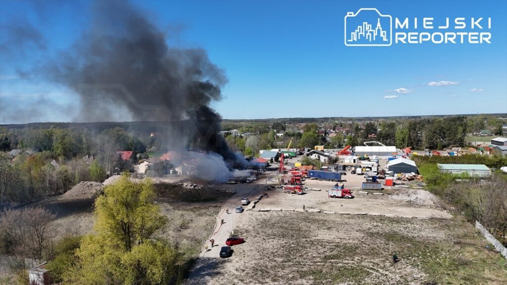 Gęsty czarny dym unoszący się nad przemysłowym terenem, w tle widać samochody strażackie i grupę ludzi.