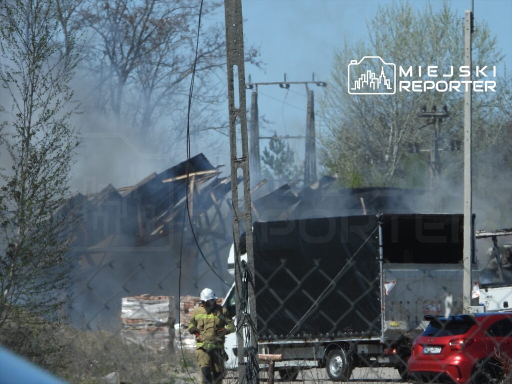 Strażak w hełmie przeszukuje teren pożaru w zrujnowanym budynku, otoczonym dymem i drewnianymi belkami.