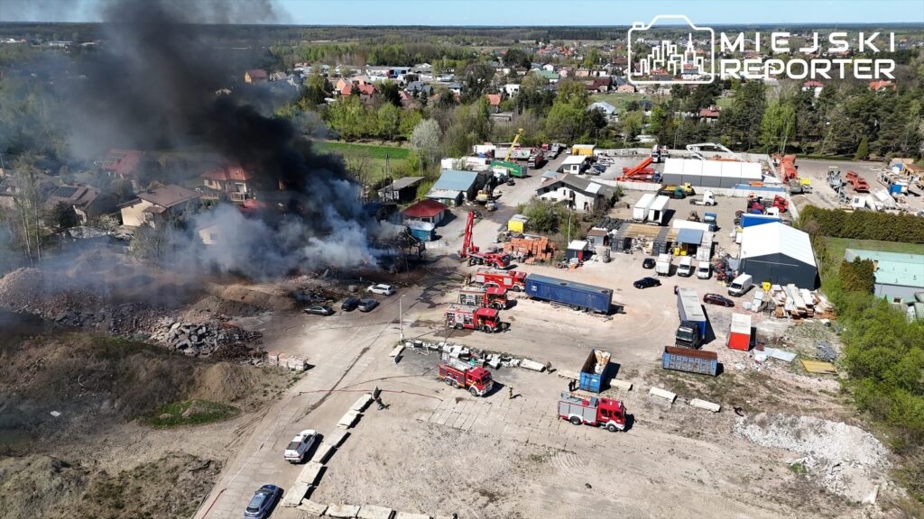 Pożar w terenie przemysłowym, z dużymi kłębami czarnego dymu unoszącymi się nad zniszczonymi budynkami oraz wozami strażackimi w akcji.