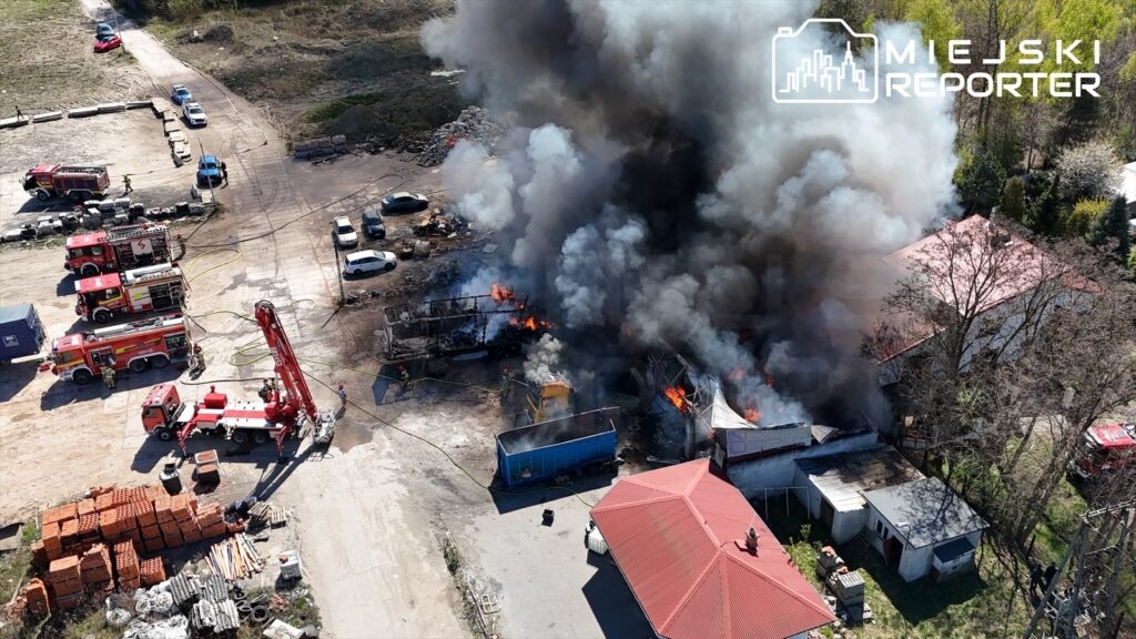 Wielka chmura czarnego dymu unosząca się nad płonącymi budynkami w strefie przemysłowej, obok pojazdy straży pożarnej.