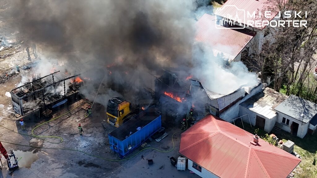 Pożar w budynku przemysłowym, gęsty dym i płomienie, strażacy gaszą ogień, w tle żółty samochód ciężarowy.