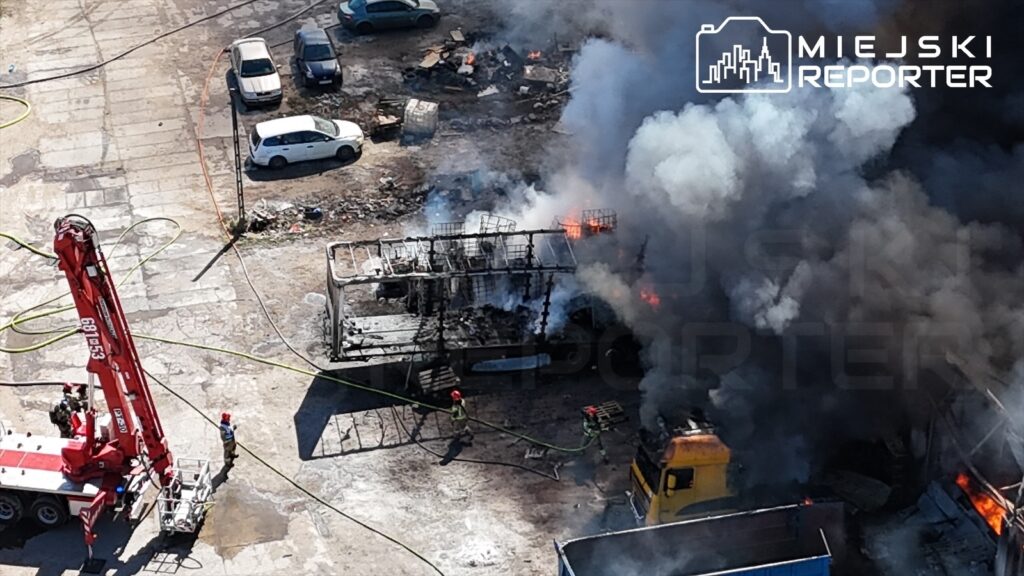 Strażacy gaszą pożar w zniszczonym magazynie, otoczonym dymem i płomieniami, w tle stoją samochody osobowe.