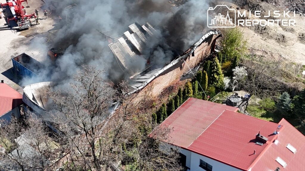 Wielka chmura dymu unosząca się z płonącego budynku w otoczeniu drzew i sąsiednich obiektów.