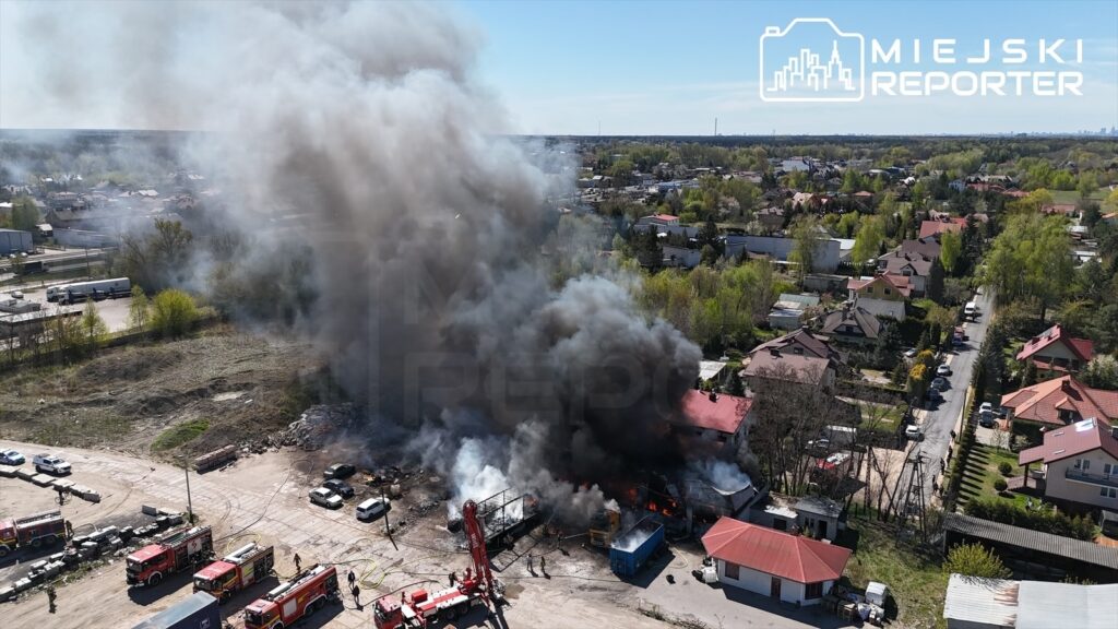 Gęsty czarny dym unoszący się z płonącego budynku w obszarze przemysłowym, wokół strażacy i pojazdy gaśnicze.