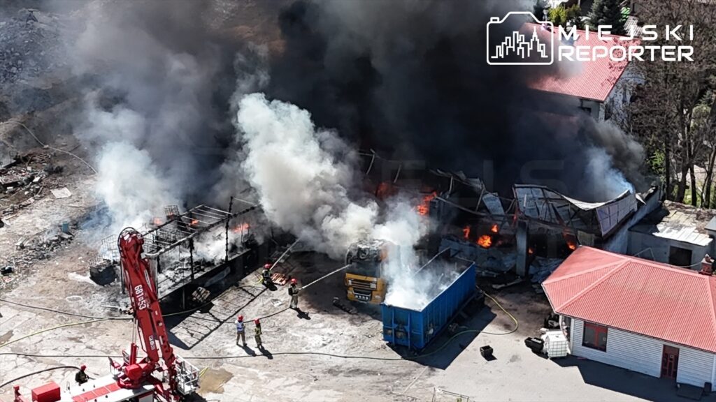 Strażacy gaszą pożar w zniszczonym budynku, otoczonym gęstym dymem, w tle widać niebieski kontener.