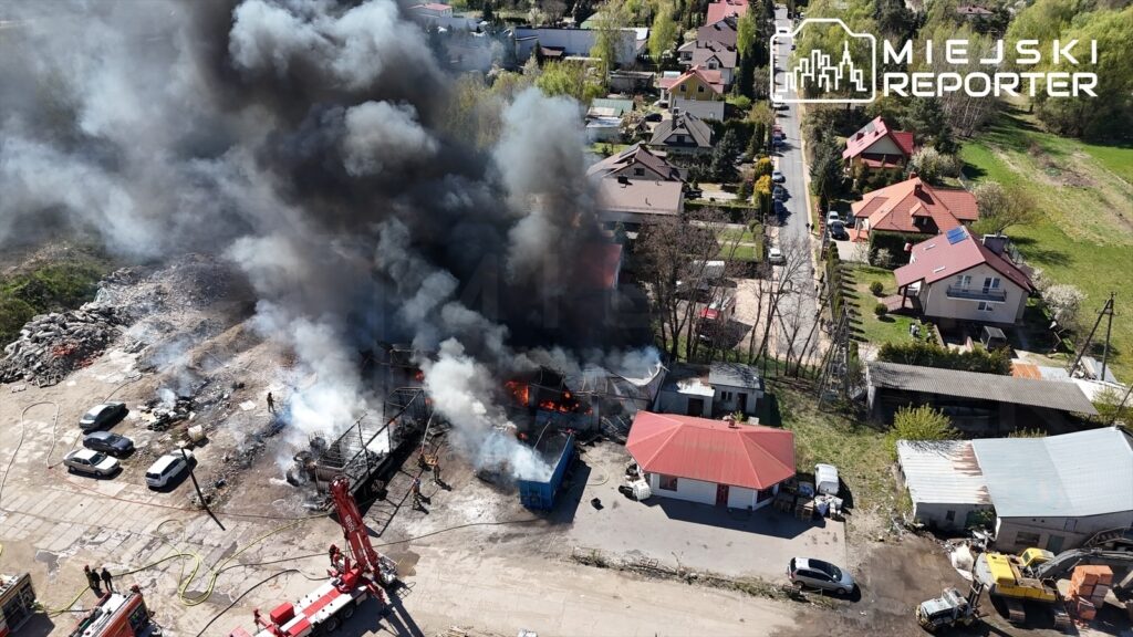 Gęsty czarny dym unoszący się z płonącego budynku w terenie przemysłowym, w otoczeniu samochodów i domów.