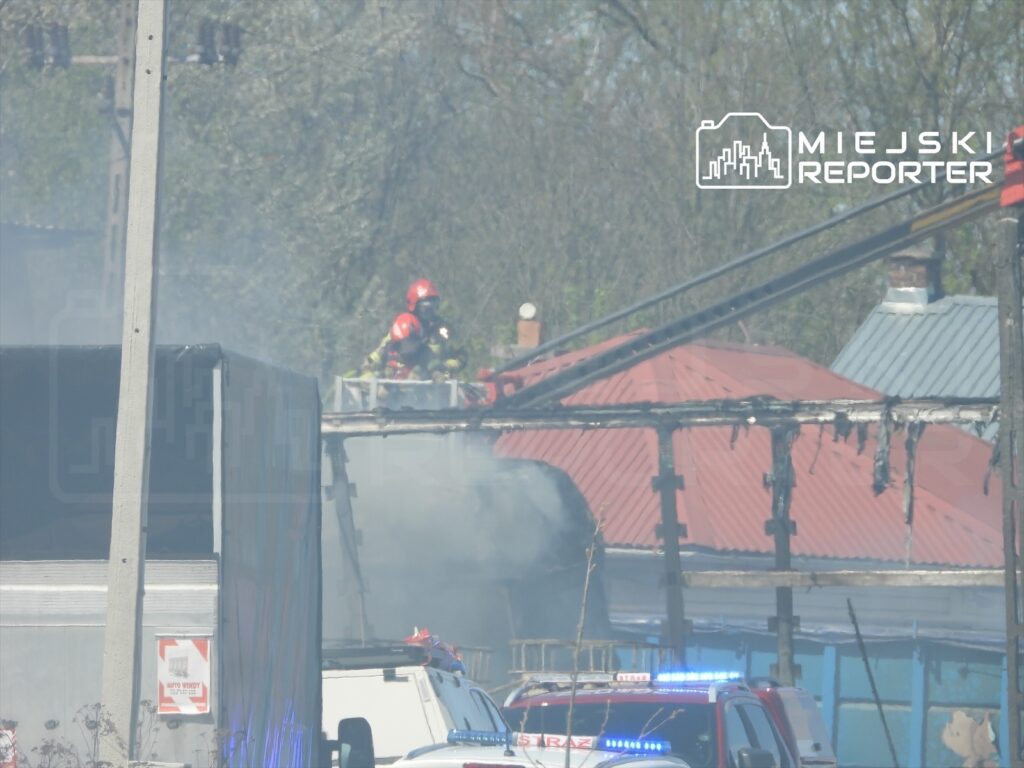 Strażacy w hełmach gaszą pożar na dachu budynku, w tle widoczne są unoszące się kłęby dymu.
