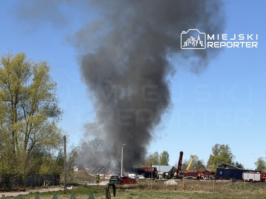Czarny dym unoszący się nad terenami przemysłowymi, w tle widoczne pojazdy strażackie i drzewa.