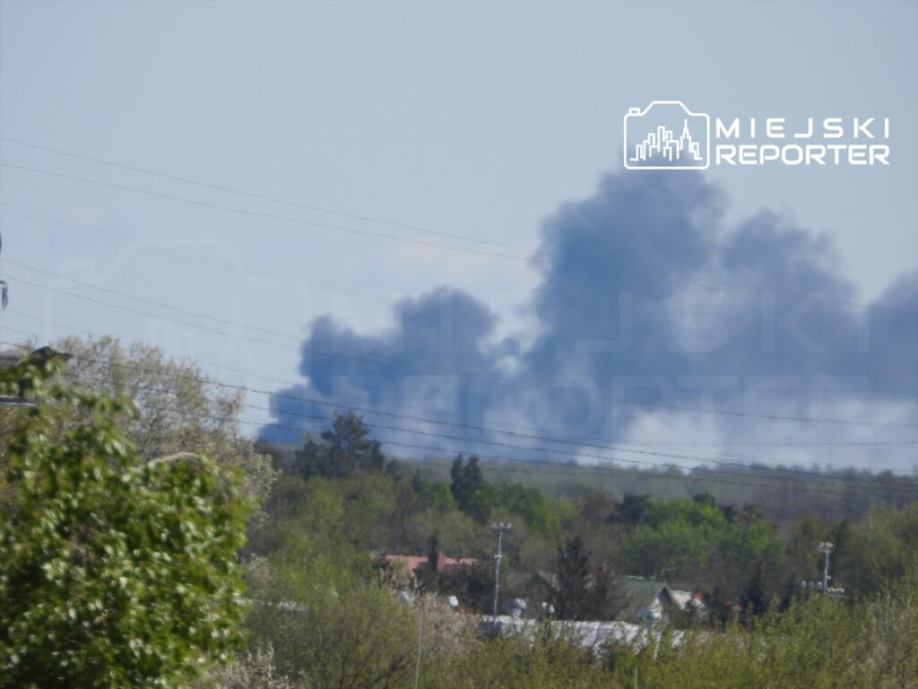 Czarny dym unoszący się nad drzewami w tle, w otoczeniu zabudowań mieszkalnych i linii energetycznych.