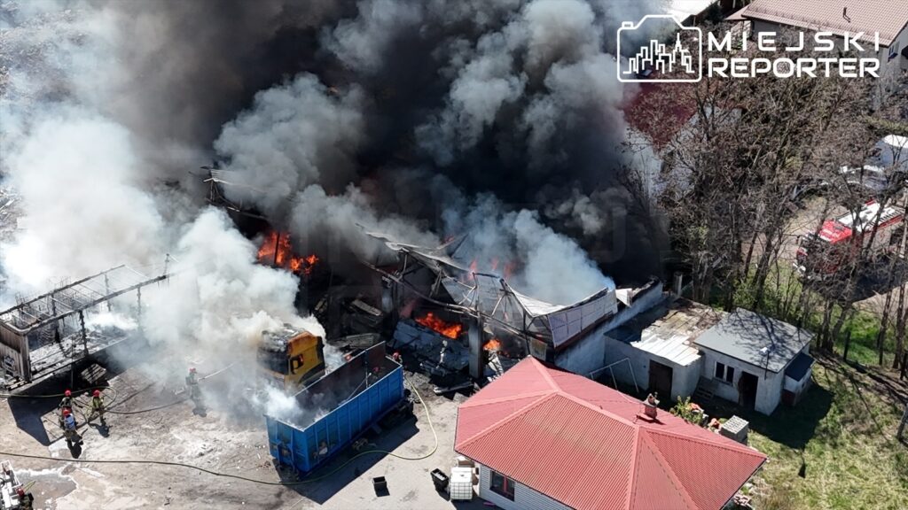 Pożar budynku z płomieniami i gęstym dymem w otoczeniu strażaków gaszących ogień. W tle widoczny czerwony dach.