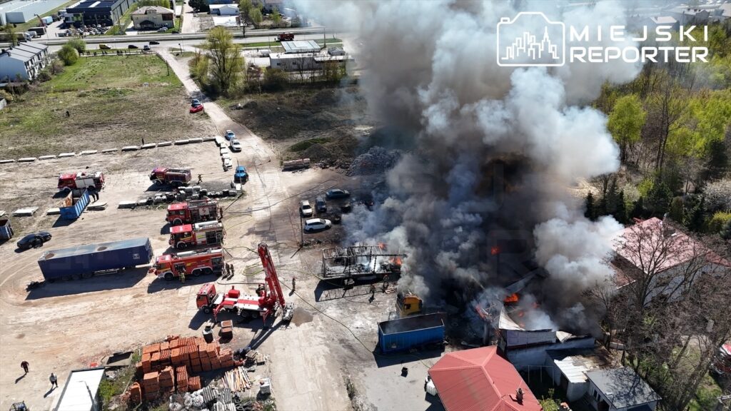 Wielka chmura czarnego dymu unosząca się nad pożarem, w którym strażacy gaszą ogień na terenie przemysłowym.