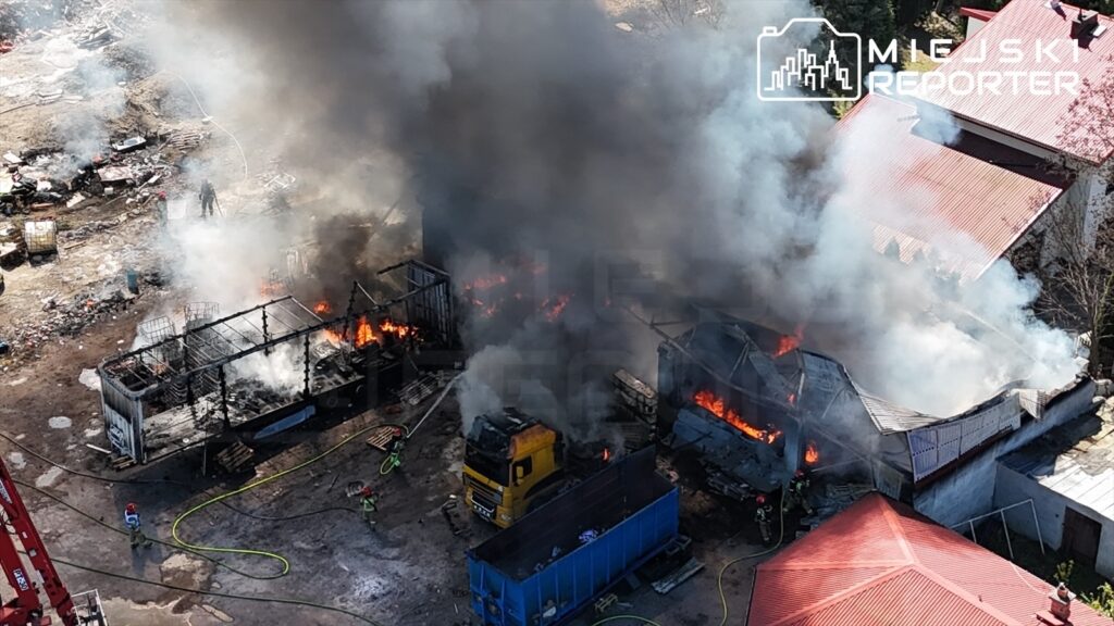 Pożar w zakładzie przemysłowym, gdzie płoną zniszczone budynki i ciężarówka; strażacy gaszą ogień w gęstym dymie.