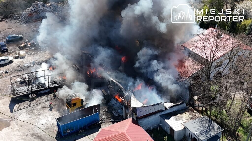 Pożar w zabudowaniach przemysłowych, z intensywnym dymem i płomieniami wydobywającymi się z budynku.