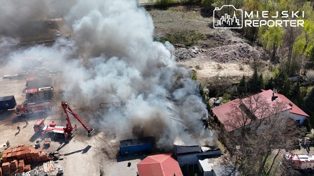 Strażacy z podnośnikiem gaszą pożar w zakładzie, otoczonym gęstym dymem, w tle widoczne budynki i sprzęt budowlany.