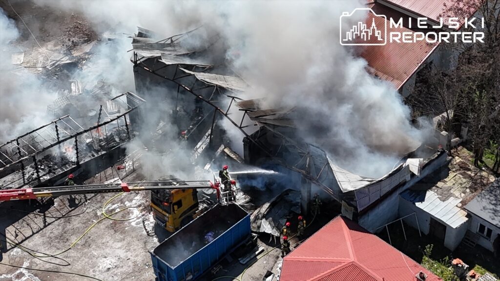 Strażacy gaszą pożar w zniszczonym budynku, otoczeni gęstym dymem i popiołem, w tle widoczne zniszczone konstrukcje.