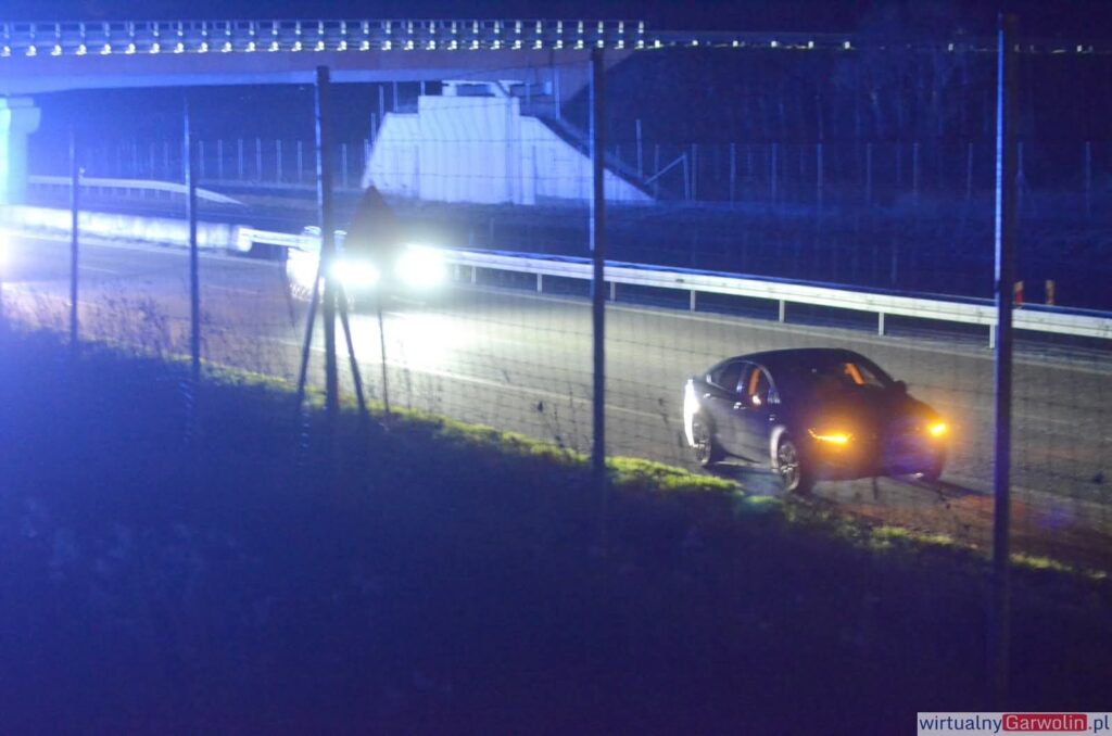 Czarny samochód zaparkowany na poboczu drogi przy oświetlonej jezdni, w tle widoczne inne pojazdy.