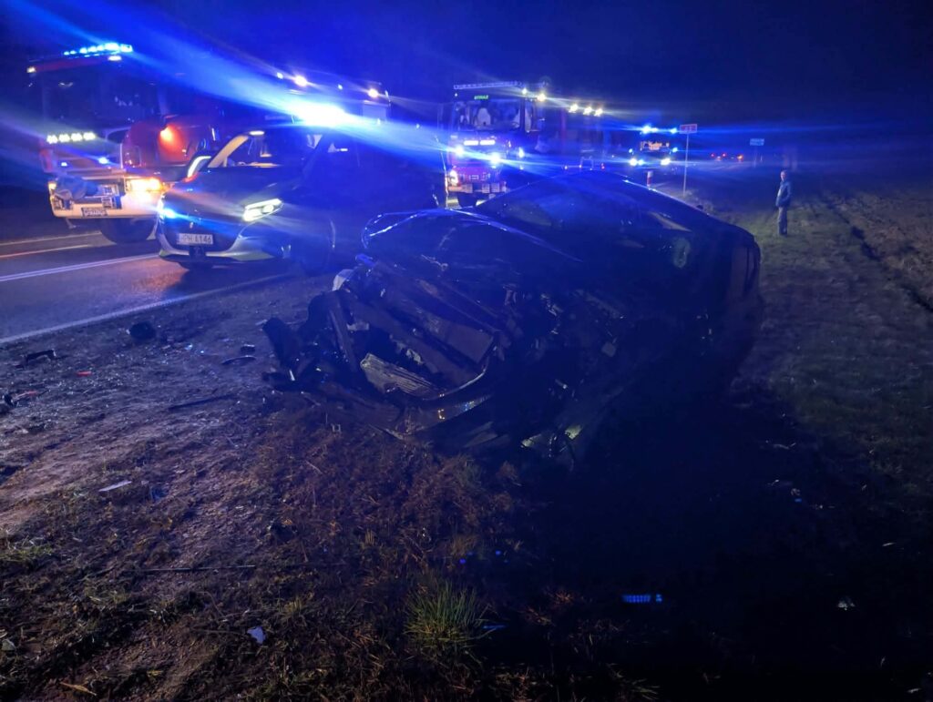 Uszkodzony samochód leży na poboczu drogi, obok pojazdy służb ratunkowych z włączonymi sygnałami świetlnymi.