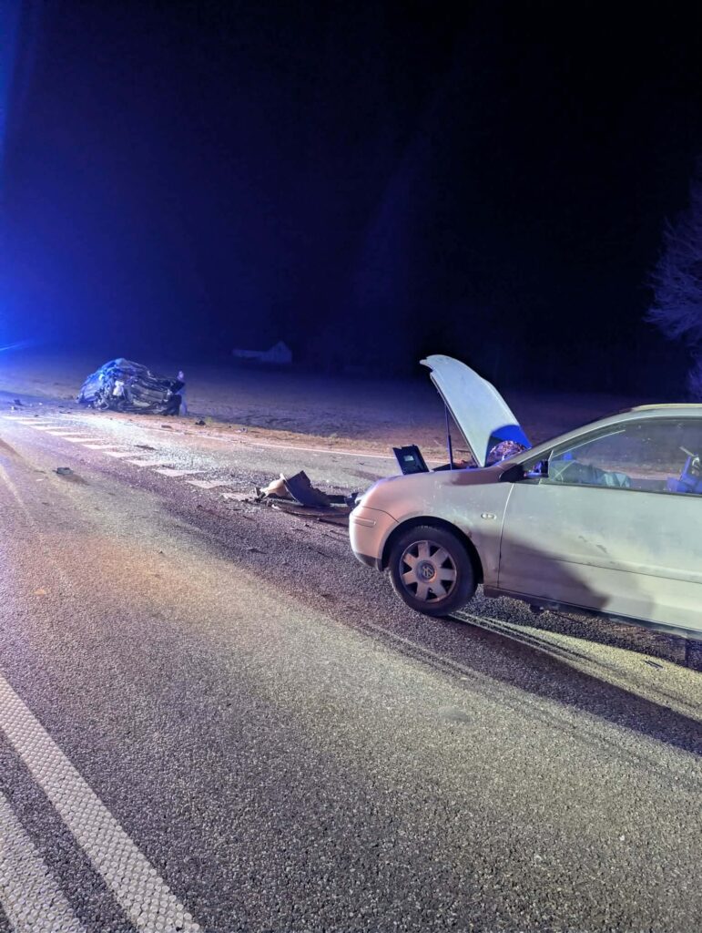 Na drodze leży rozbity samochód, a obok stoi inny pojazd z otwartą maską. W tle widoczne światła policyjne.