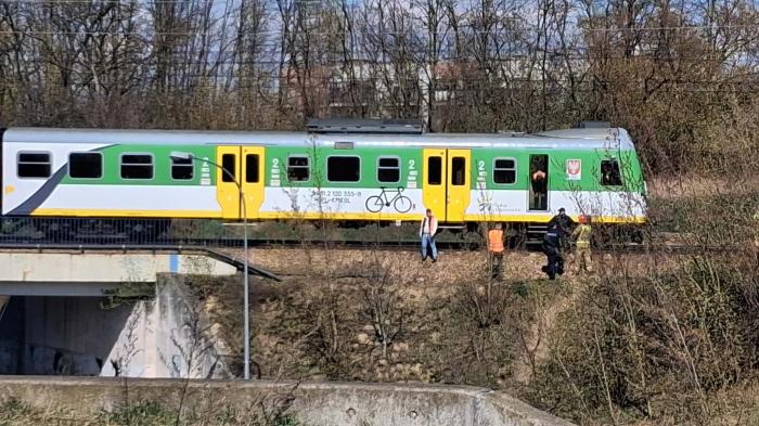 Pociąg osobowy zatrzymany na torach, obok grupy osób w odzieży roboczej oraz pasażerów na peronie.